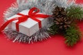 Christmas card. White gift box tied with a red bow with Christmas tree branches, pine cones and silver mesh on a red background.