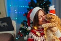 Christmas celebration with cat. Woman playing and kissing pet in Santa`s hat by New year tree at home Royalty Free Stock Photo