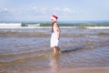 Happy cute teenage girl in a red Santa hat sitting on a sand on a sea beach and making a date 2020 from shells, vertical