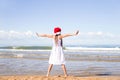 Happy little girl wearing a red Santa hat standing arms outstretched on the sandy beach by the sea on clear sunny day Royalty Free Stock Photo