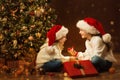 Christmas Child Girl opening Box and giving Gift Toy to Sister. Happy Children in Santa Hat next to Xmas Tree. Fairy Magic