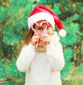 Christmas child little girl in santa red hat playing with sweet lollipop cane near tree