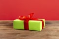 Christmas composition. Green gift box with red ribbon on a wooden table and red background. Side view. a Christmas gift