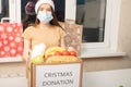 Christmas covid donation. A young female volunteer wearing a medical mask at a charity center collects food food in a box for