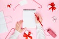 Christmas cozy background. Female hand writing in blank notebook with festive decorations, gift boxes on pastel pink. New year Royalty Free Stock Photo