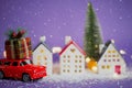 Christmas decor - red retro car on snow carries past houses with fairy lights in bokeh Christmas tree with gift boxes on roof. Toy Royalty Free Stock Photo