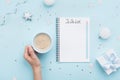 Christmas do list and woman hand with cup of coffee on blue pastel table top view. Flat lay style. Holiday planning.