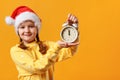 Christmas Eve. Portrait of a Cute little girl in a Santa hat. The child is holding an alarm clock on a yellow background Royalty Free Stock Photo