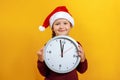 Christmas Eve. Portrait of a happy charming cute little girl in Santa`s hat. The child holds a clock on a yellow background Royalty Free Stock Photo