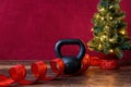 Christmas fitness, black kettle bell, with artificial Christmas tree and white lights, red ribbon, wood table, red background Royalty Free Stock Photo