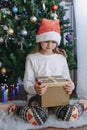 Christmas gifts for children. A cute Caucasian girl in a New Year`s hat opens a box with a gift under a New Year tree. Vertical Royalty Free Stock Photo