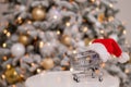 Christmas gifts shopping time. Shopping trolley in a santa claus hat by the christmas tree. Royalty Free Stock Photo