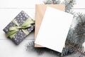 Christmas greeting card mockup with craft paper envelope, green color gift box and fresh fir tree branch on white wooden