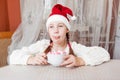 baby girl in Santa hat sits at the table and holds a large mug of hot chocolate and marshmallows. Royalty Free Stock Photo