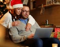 Christmas holiday. Female in Santa`s hat hugging a man using a laptop. Royalty Free Stock Photo