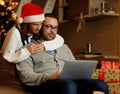 Christmas holiday. Female in Santa`s hat hugging a man using a laptop. Royalty Free Stock Photo