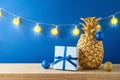 Christmas holiday greeting card with pineapple as alternative Christmas tree, lights garland and gift box on wooden shelf over