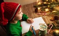 Christmas Kid writing Letter to Santa. Happy Child in Red Santa Hat dreaming about Presents at Home. Xmas Gifts Shopping Wish List