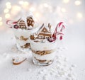 Christmas layered cream and gingerbread dessert decorated with gingerbread houses and candy cane in glasses on white table