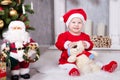 Christmas or New year celebration. Little girl in red dress and santa hat with bear toy sitting on the floor near the Christmas tr Royalty Free Stock Photo