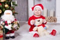 Christmas or New year celebration. Little girl in red dress and santa hat with bear toy sitting on the floor near the Christmas tr Royalty Free Stock Photo