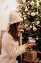 Christmas, New Year. Pretty young woman in white sweater and knitted hat with luminous gift box near christmas tree, new year m