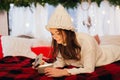 Christmas, New Year. Pretty young woman in white sweater and knitted hat lying in bed with luminous gift box, new year magic atm