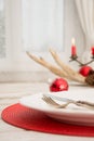 Christmas place setting with white dishware, silverware and red decorations on white board in interior near the window. Royalty Free Stock Photo