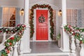 Christmas porch decoration idea. House entrance with red door decorated for holidays. Red and green wreath garland of fir tree