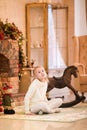 Christmas portrait of happy blonde child girl in white sweater siting on the floor near the Christmas tree and wooden toy horse. N Royalty Free Stock Photo