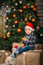 Christmas portrait of happy smiling little boy in red santa hat sitting on boxes with presents holding oranges in hands Royalty Free Stock Photo