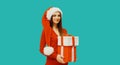 Christmas portrait of happy smiling woman holding gift box in her hands wearing red santa claus hat on blue studio background Royalty Free Stock Photo