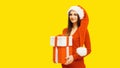 Christmas portrait of happy smiling woman holding gift box in her hands wearing red santa claus hat on yellow background Royalty Free Stock Photo