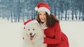 Christmas portrait of happy woman with white Samoyed dog in red santa hat sitting on snow in winter Royalty Free Stock Photo