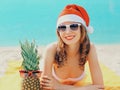 Christmas portrait of happy young smiling woman in red santa hat with funny pineapple lying on a beach together over sea