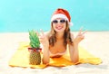 Christmas portrait pretty young smiling woman in red santa hat and pineapple lying on beach over sea