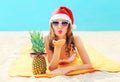 Christmas portrait pretty young woman in red santa hat with pineapple lying on beach sends air kiss over blue sea Royalty Free Stock Photo