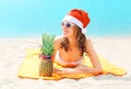 Christmas portrait pretty young woman in red santa hat with pineapple lying on beach sends air kiss over blue sea Royalty Free Stock Photo