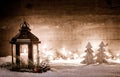 Christmas scene with a lantern, trees, fir branch, snow flakes and blurred lights in front of an illuminated dark wooden board as