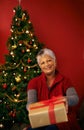 Christmas, senior and happy woman with gift in portrait at studio for holiday celebration by tree isolated on a red Royalty Free Stock Photo