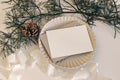 Christmas still life. Blank horizontal greeting card, invitation mockup on scalloped plate in sunlight. Pine cone, pine