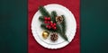Christmas table with decorations, white empty plate on red tablecloth, green background, top view, above. Celebration xmas eve: Royalty Free Stock Photo