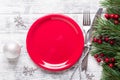 Christmas table setting with empty red plate, gift box and silverware on light wood background. Fir tree branch, holly berries.