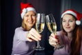 Christmas toast with glasses of champagne in hands. Blurred background: two girls in Santa Claus hats, clink glasses and wish Royalty Free Stock Photo