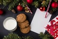 Christmas tree branches with cones and decorations on a dark table next to a gift sheet and milk with cookies