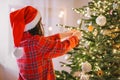 Christmas tree Decorating. Girl child in santa hat and plaid pajamas decorate the Christmas tree with red shiny balls Royalty Free Stock Photo