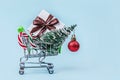 Christmas tree, gift box and candy canes in miniature shopping cart with on pastel blue background with copy space.