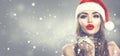 Beauty model woman in Santa`s hat blowing snow in her hand. Christmas winter fashion girl on holiday blurred winter background Royalty Free Stock Photo