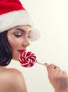 Christmas woman. Beauty model girl in Santa Claus hat eating red xmas lollipop candy. Closeup portrait on grey background Royalty Free Stock Photo