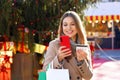 Christmas woman carrying shopping bags outdoor. Beautiful smiling girl paying her gifts online with mobile phone and credit card Royalty Free Stock Photo